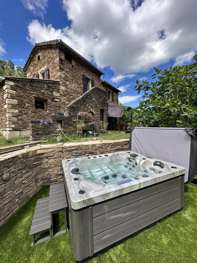 Spa extérieur luxe face à une magnifique maison de pierre. Jacuzzi moderne sur gazon synthétique devant une maison rurale en pierre naturelle et ses murs de soutènement sous un ciel bleu.