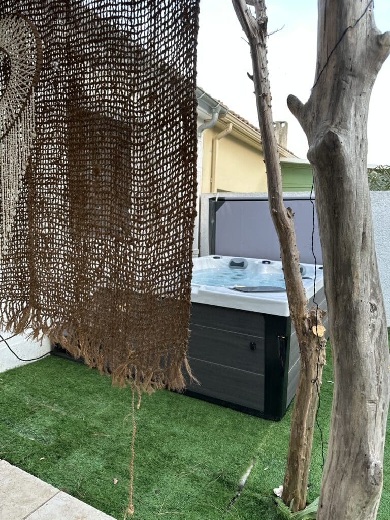 Jacuzzi extérieur moderne et jardin privé Jacuzzi extérieur gris moderne sur gazon artificiel. Le bain à remous est encadré par un filet de jute et des troncs en bois brut.