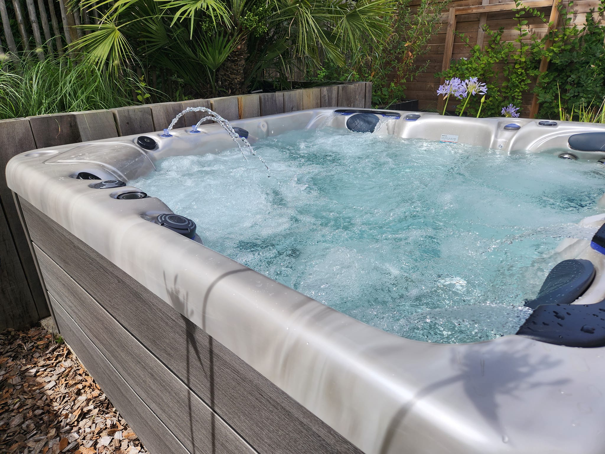 Bain à remous moderne et bouillonnant dans un jardin vert. Les fontaines animent le spa aux finitions grises.