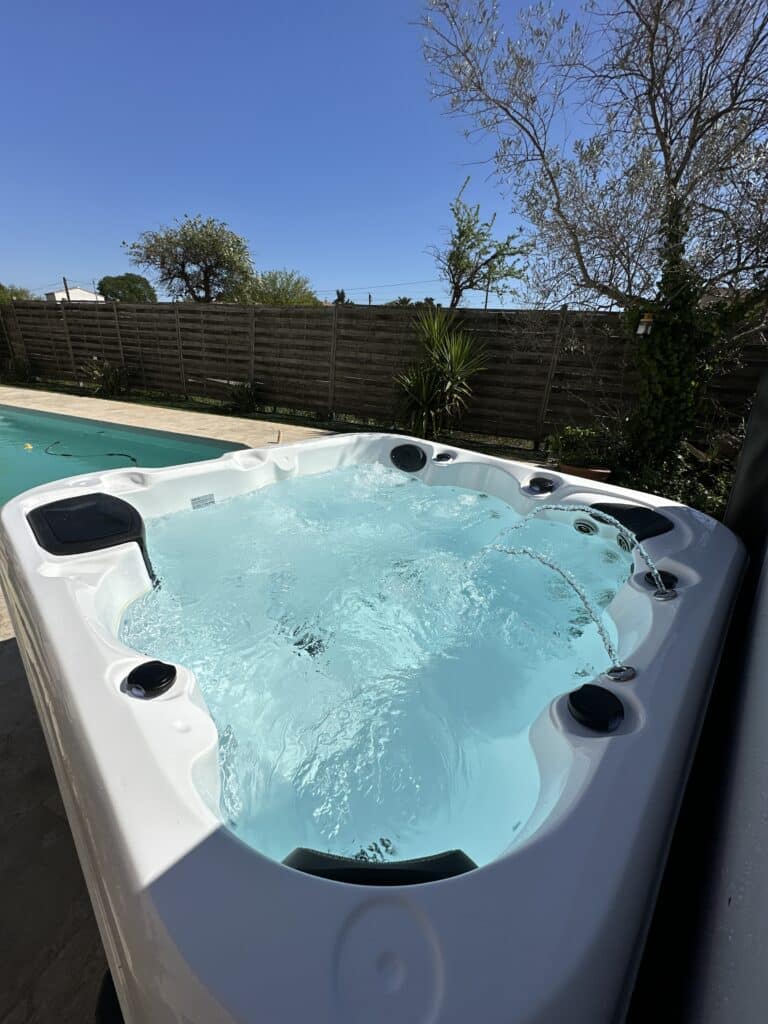 Grand jacuzzi blanc bouillonnant avec jets d'eau à côté d'une piscine extérieure ensoleillée. Ambiance détente, clôture en bois et ciel bleu.