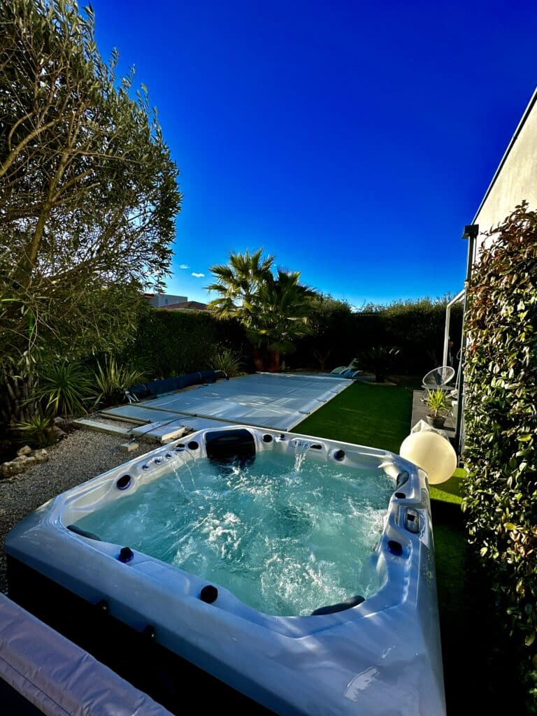 Jacuzzi extérieur bouillonnant sous un ciel bleu éclatant dans un jardin luxuriant avec un palmier et une piscine couverte.