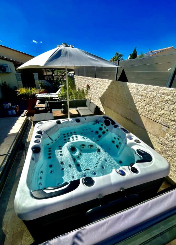 Spa extérieur moderne d'eau turquoise sur une terrasse ensoleillée. Il est entouré d'un mur de pierre, d'un parasol blanc et d'un salon de jardin.