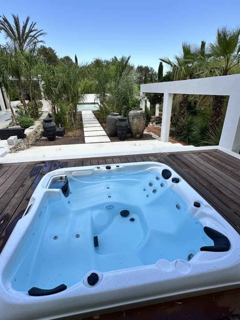 Jacuzzi bleu vif sur une terrasse en bois, avec un jardin tropical luxuriant, des palmiers, des jarres antiques et un chemin menant à la piscine.