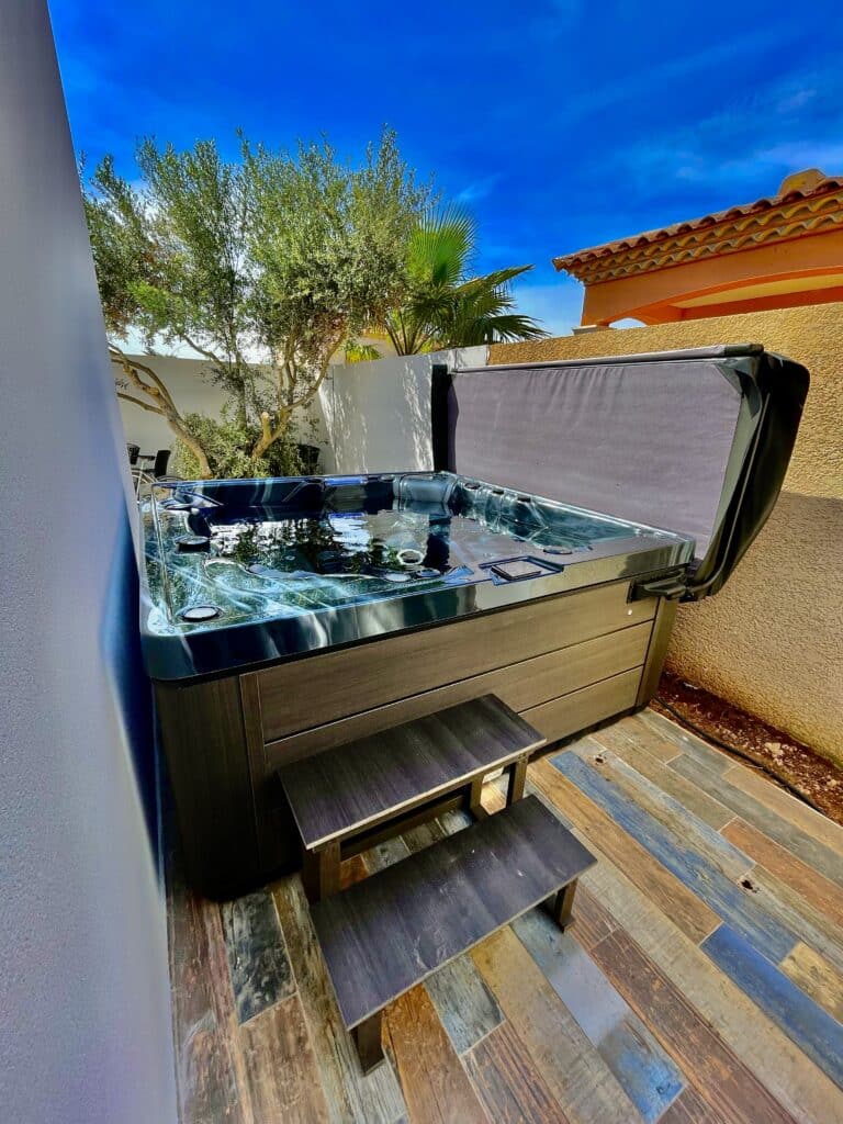 Spa extérieur de luxe : profitez du jacuzzi sur la terrasse. Jacuzzi extérieur moderne sur une terrasse colorée style bois patiné, entouré d'arbres et d'un mur blanc, sous un ciel bleu azur.