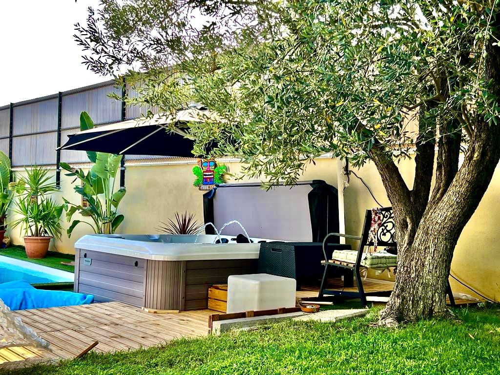 Jacuzzi et espace détente : luxe et fraîcheur au jardin. Grand jacuzzi extérieur gris sur une terrasse bois. L'espace détente est ombragé par un olivier et un parasol, dans un jardin luxuriant.