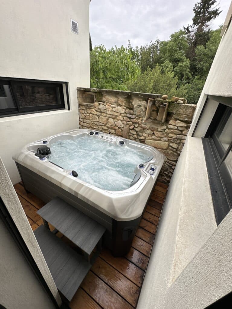 Jacuzzi privé extérieur : luxe et détente sur terrasse bois. Bain à remous extérieur bouillonnant sur une terrasse en bois. Murs blancs modernes, escaliers gris, et mur rustique en pierres sous les arbres.