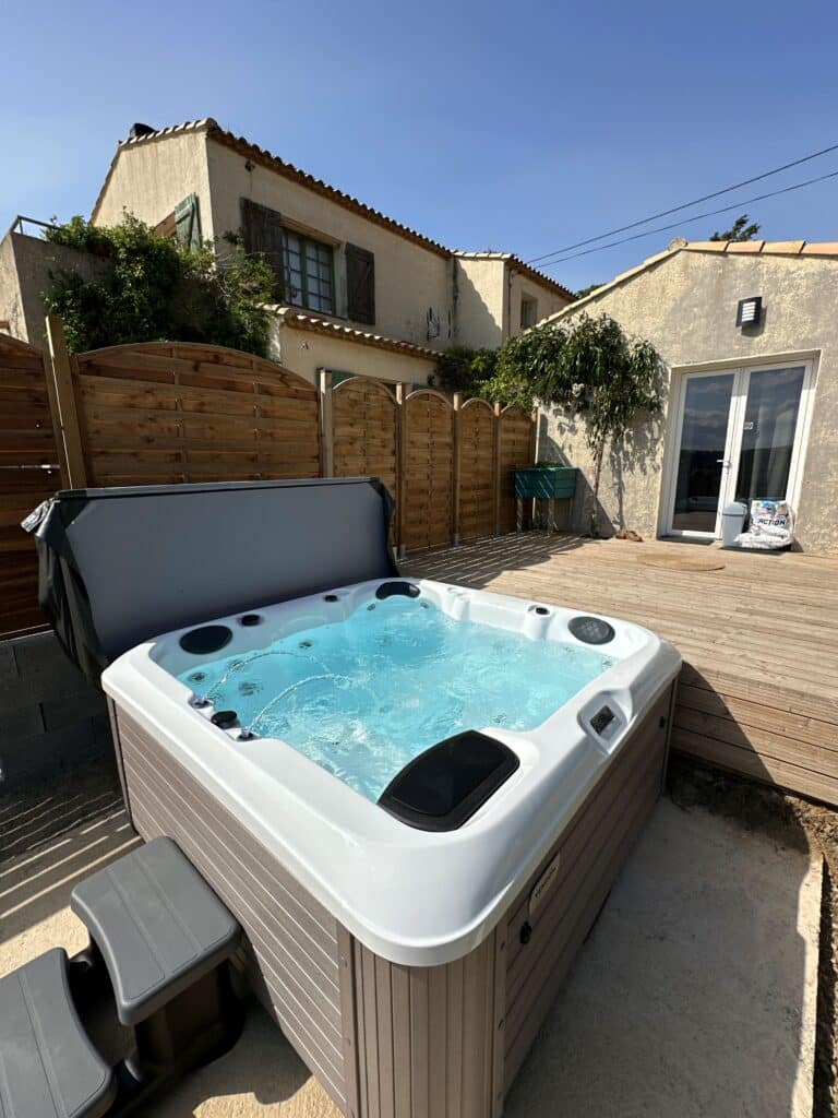 Jacuzzi extérieur bouillonnant sur terrasse ensoleillée Jacuzzi moderne avec eau turquoise bouillonnante installé sur une terrasse en bois. Façade de maison ensoleillée avec palissade.