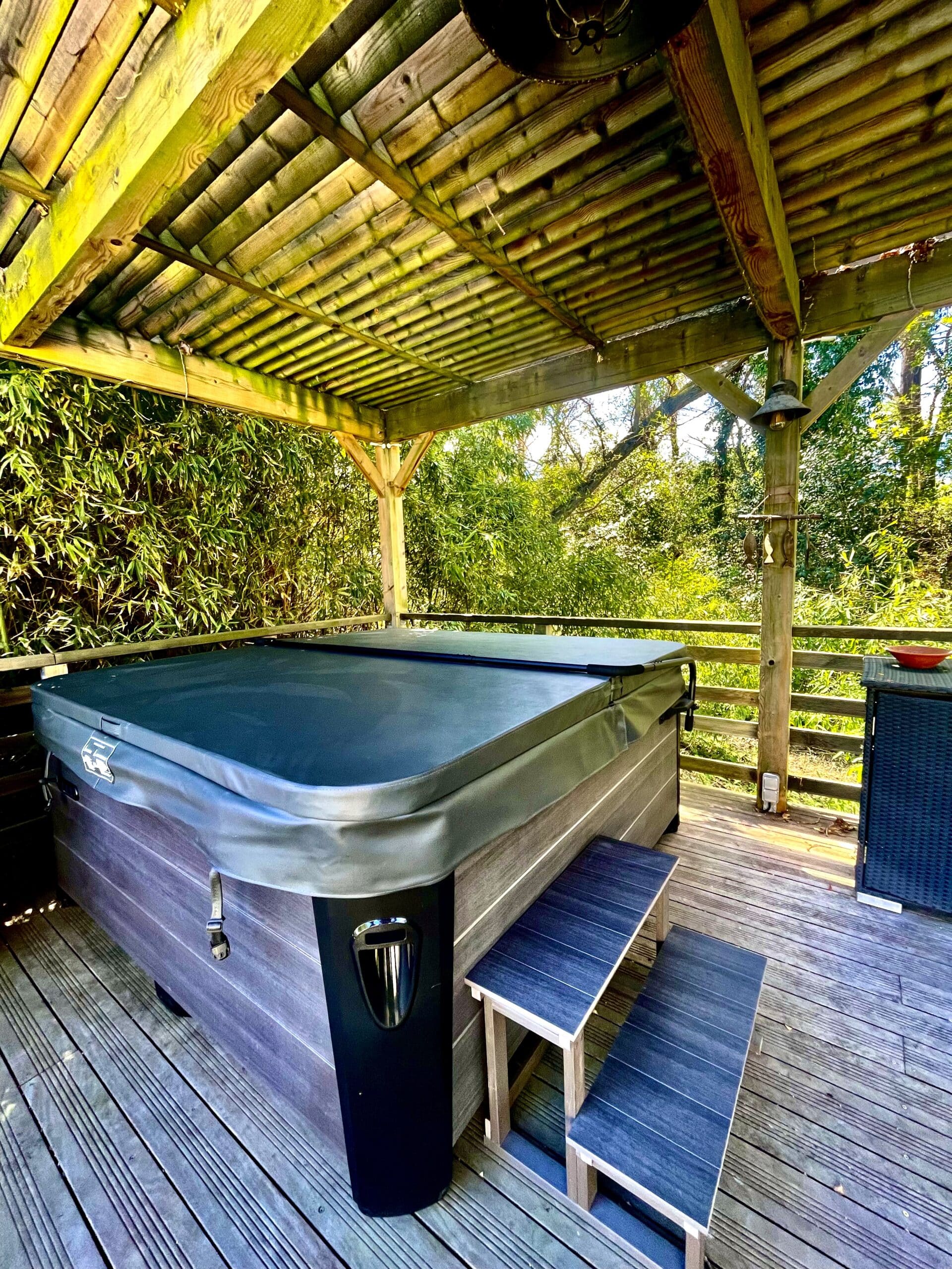Jacuzzi extérieur moderne sur terrasse en bois, couvert par une pergola rustique. Environnement boisé dense et intimiste.