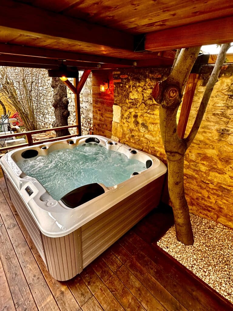 Spa extérieur bouillonnant sur terrasse en bois. Mur de pierre rustique et éclairage ambré sous une pergola créent une ambiance chaleureuse.