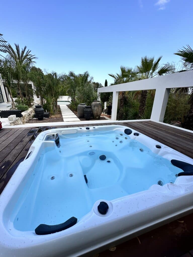 Jacuzzi extérieur sur terrasse bois pour une détente luxueuse Jacuzzi blanc et bleu intégré à une terrasse en bois sombre, entouré de palmiers et d'un jardin luxuriant de style méditerranéen.