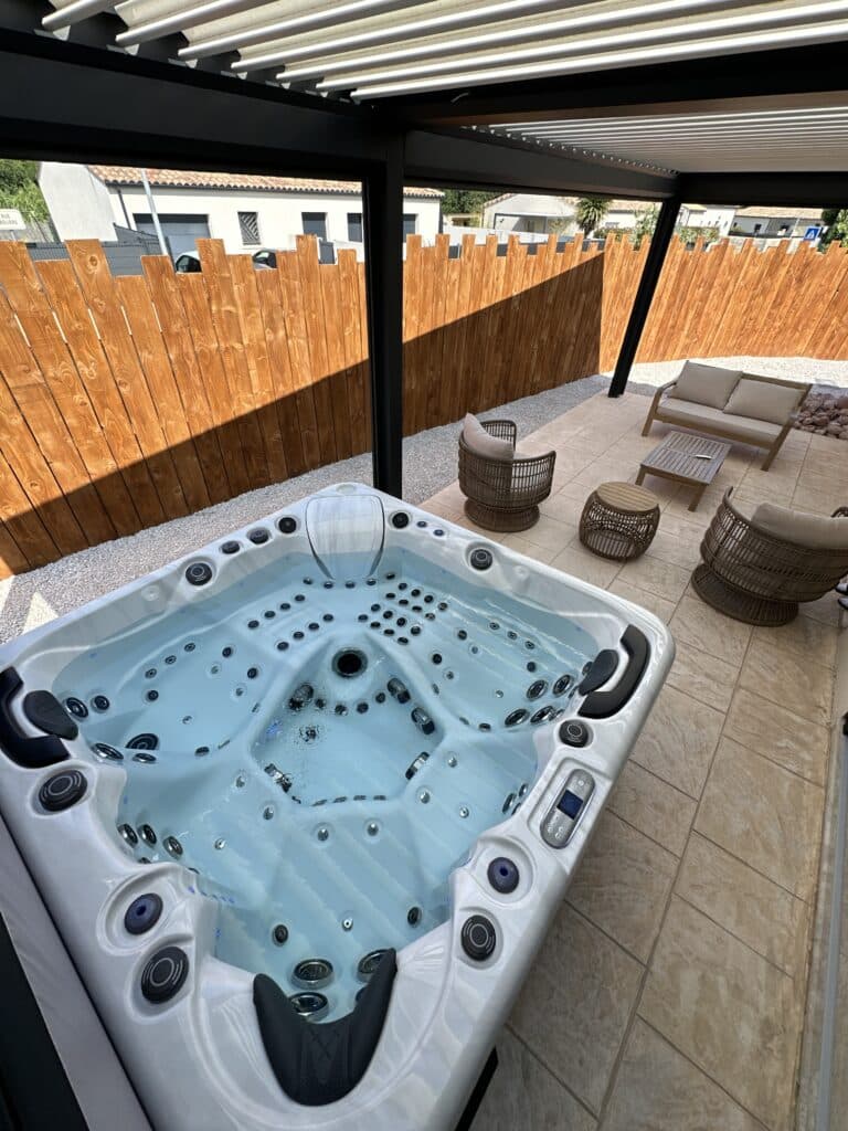 Luxueux jacuzzi blanc sur une terrasse ensoleillée, entouré de mobilier de jardin et d'une clôture en bois, sous une pergola.