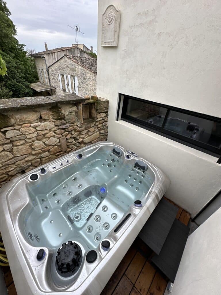 Spa extérieur bouillonnant sur terrasse en bois, encadré par un mur de pierre rustique et un mur moderne affichant "CARPE DIEM".