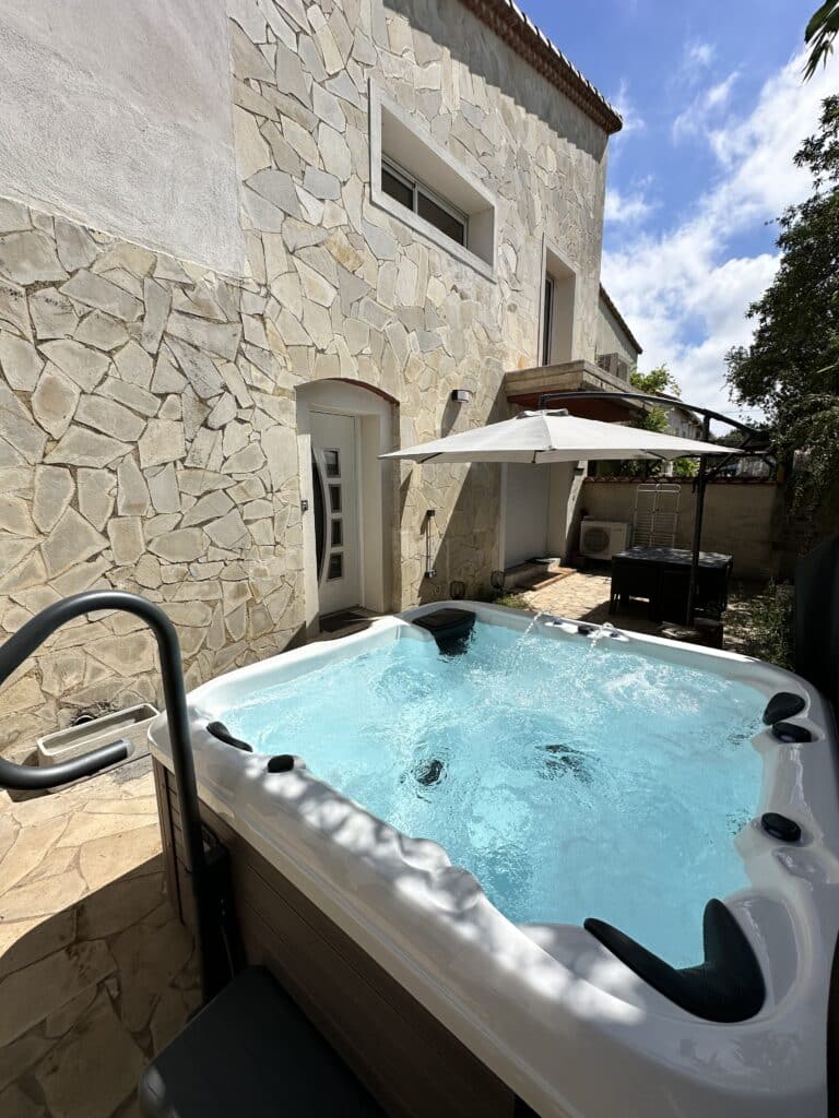 Jacuzzi extérieur privé sur terrasse en pierres claires. Jacuzzi extérieur bouillonnant sur une terrasse pavée, adossé à une façade en pierres de taille claires, sous un ciel bleu.