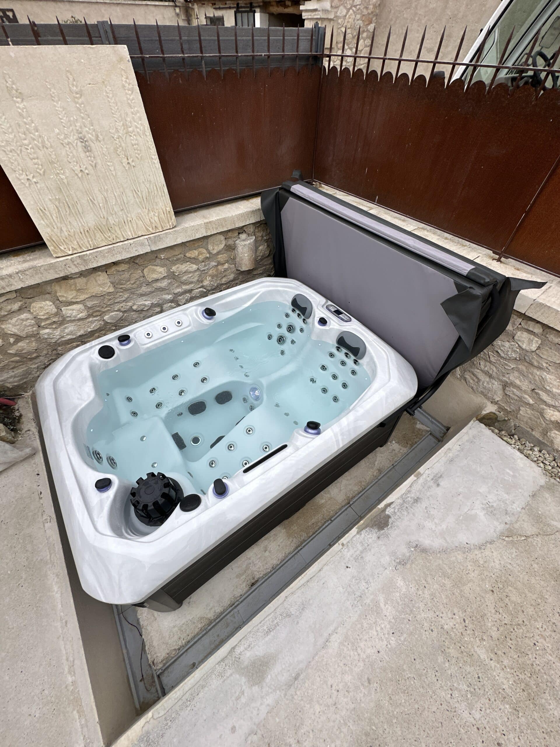 Bain à remous blanc extérieur semi-enterré sur terrasse en béton, près d'un mur de pierre et d'une clôture en métal rouillé.