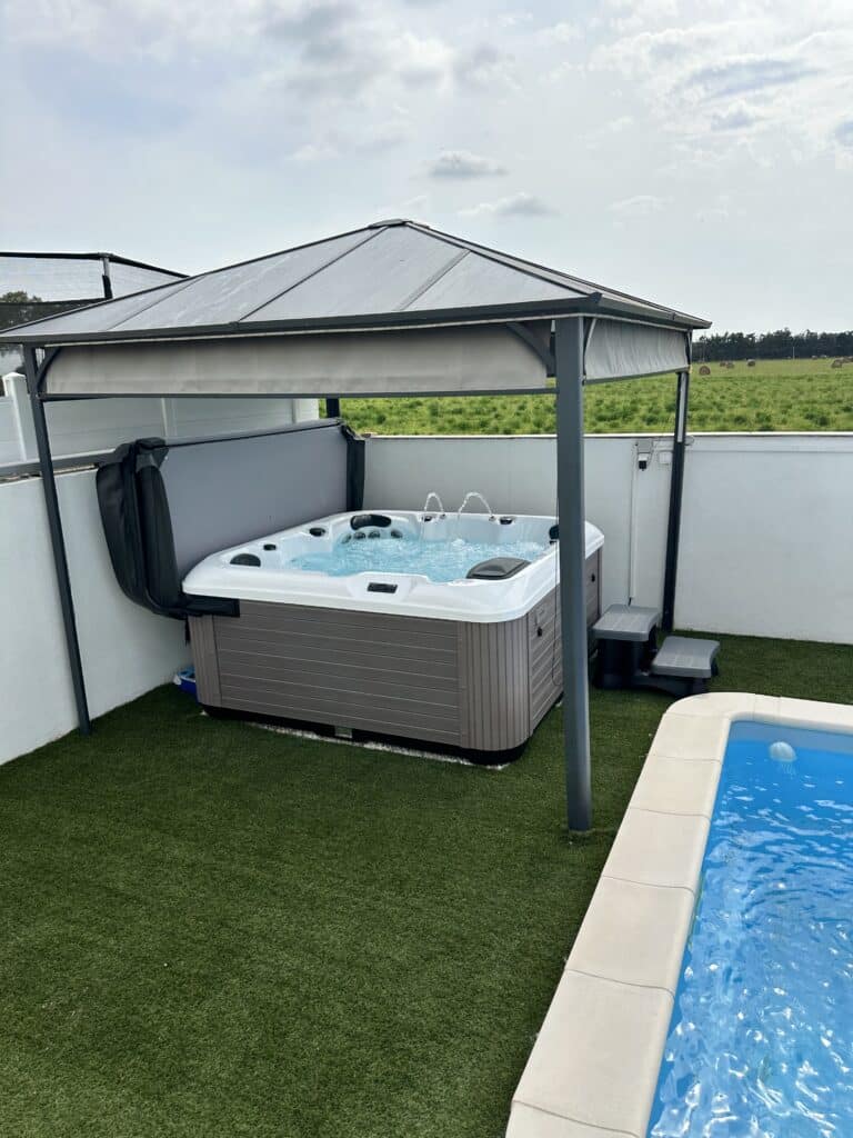 Spa extérieur sous tonnelle, vue sur champ de foin. Jacuzzi extérieur gris sous abri, avec jets d'eau, installé sur gazon synthétique près d'une piscine et d'un champ de foin.