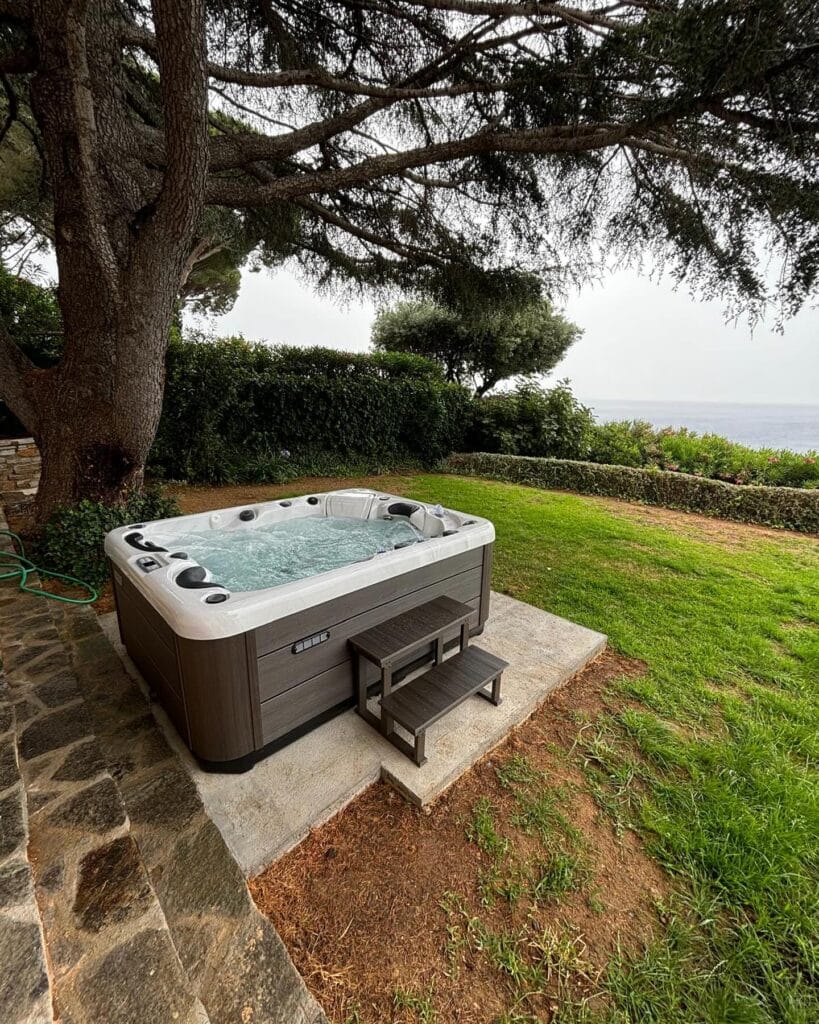 Jacuzzi extérieur vue mer : le spa de luxe pour votre jardin. Spa extérieur moderne sous un grand pin, avec marches en bois composite, installé sur un patio en pierre et offrant une vue imprenable sur la mer.
