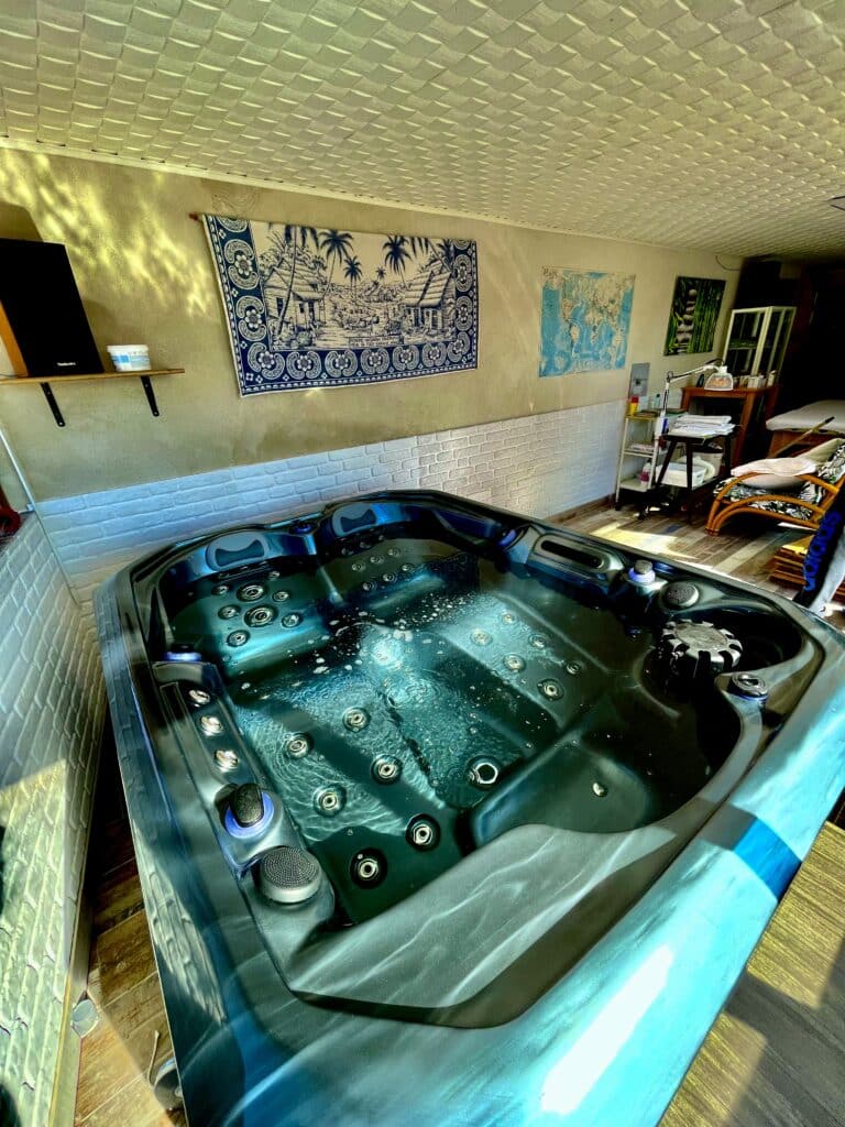 Détente maximale : Jacuzzi intérieur et salle de spa privée. Jacuzzi intérieur bleu moderne bouillonnant. Salle de spa/détente avec murs briques, tenture tropicale bleue et équipements de massage.