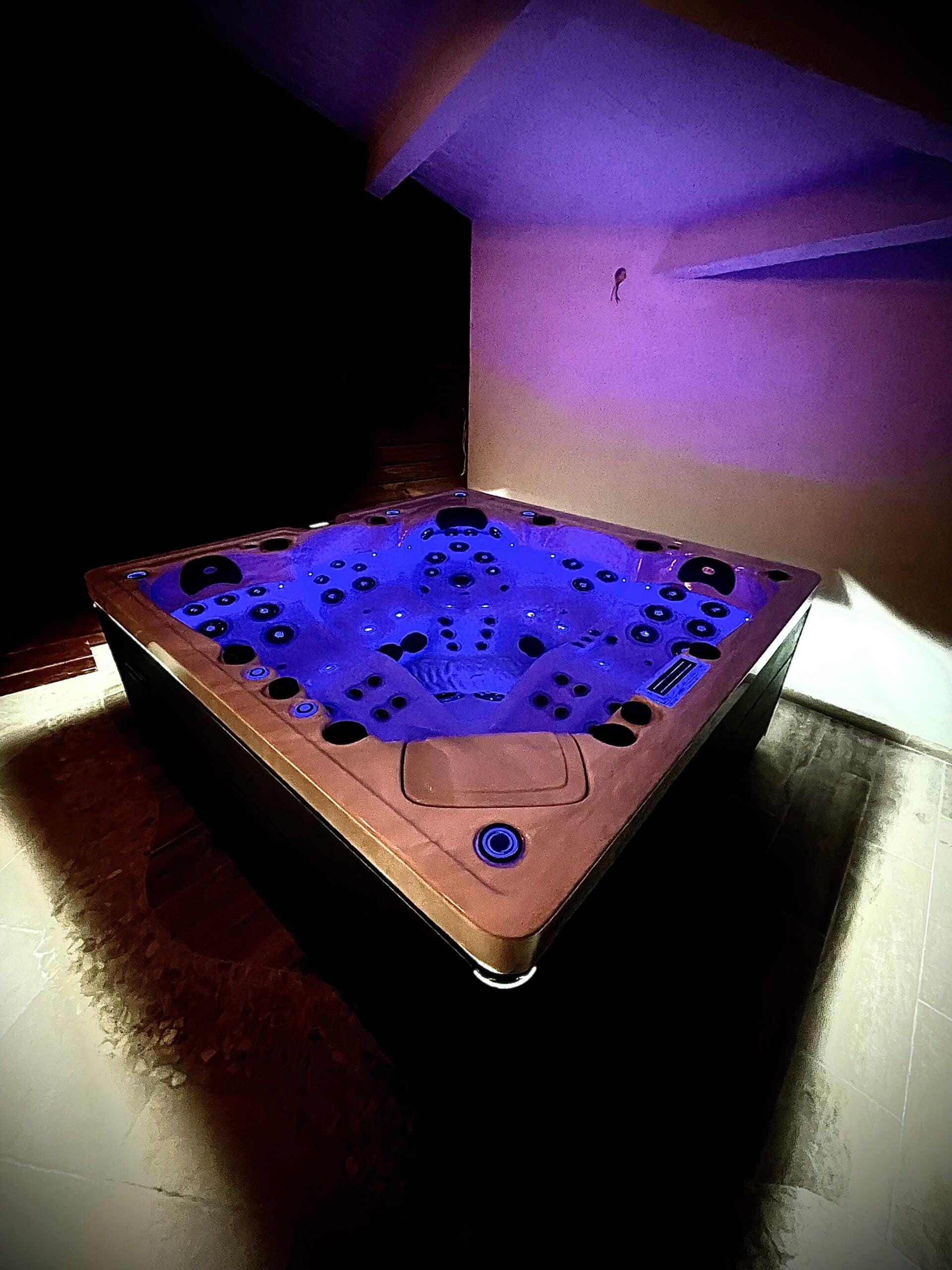 Jacuzzi intérieur moderne éclairé par une intense lumière LED bleue et violette dans une pièce sombre, invitant à la détente.