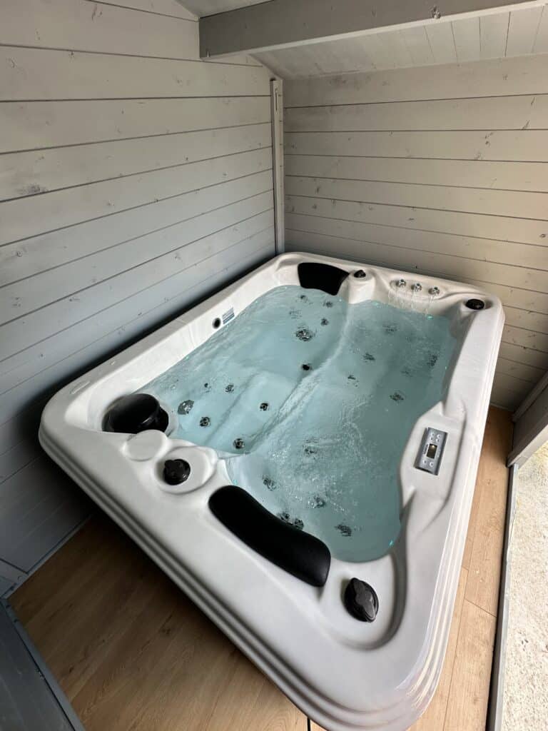 Jacuzzi blanc bouillonnant (39.0°C) dans un abri privé en bois gris clair. Murs lambrissés et plancher en bois brun.
