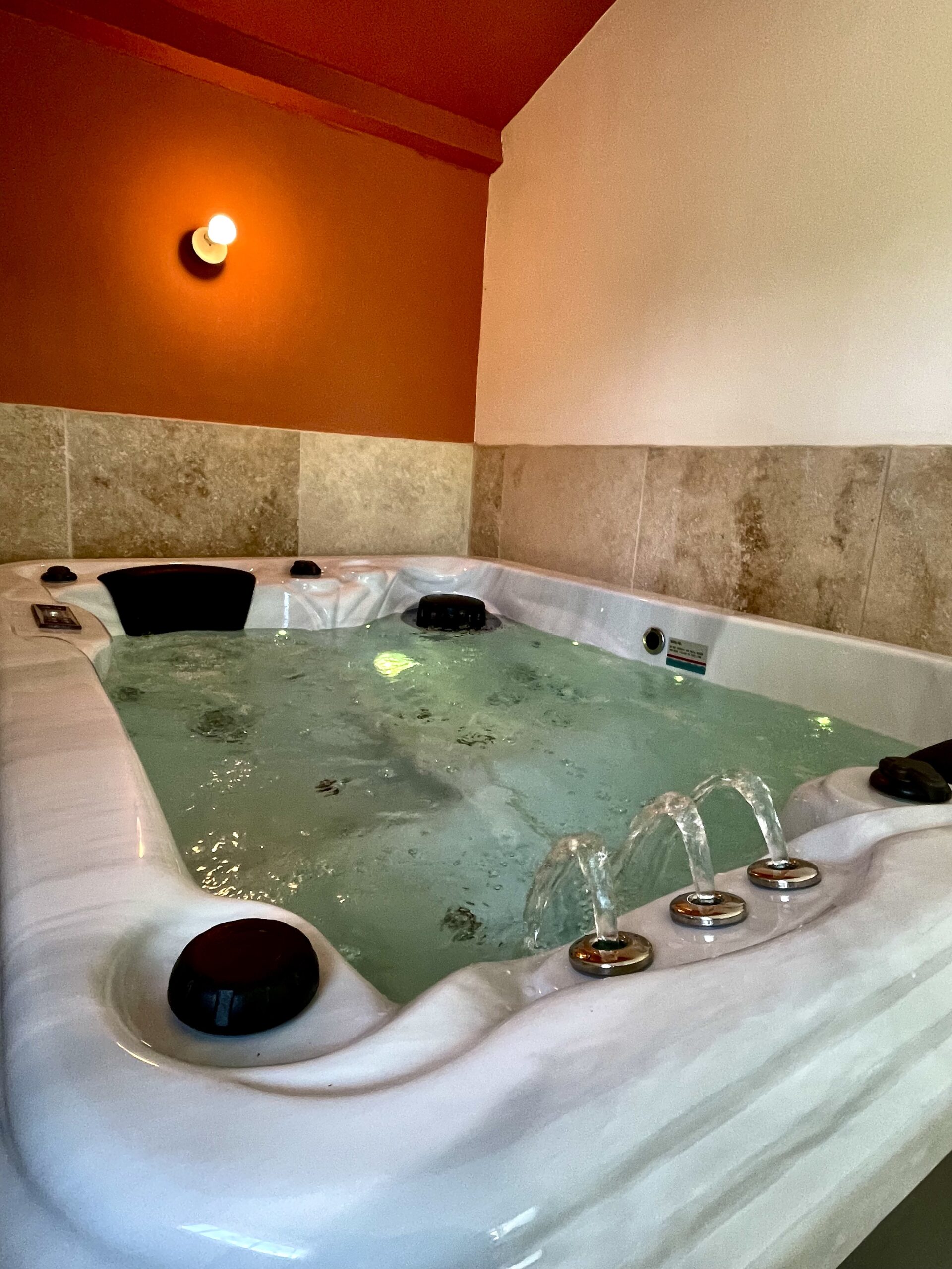 Jacuzzi intérieur bouillonnant avec fontaines, sur fond de mur orange terracotta et de carrelage en pierre beige. Ambiance lumière chaude.