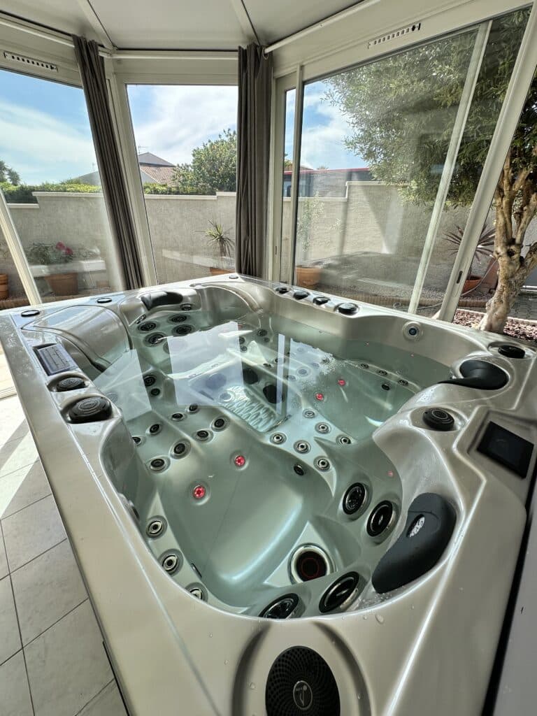 Jacuzzi intérieur moderne dans une véranda lumineuse. Bain à remous aux jets rouges allumés, prêt pour la détente. Vue sur jardin privé.