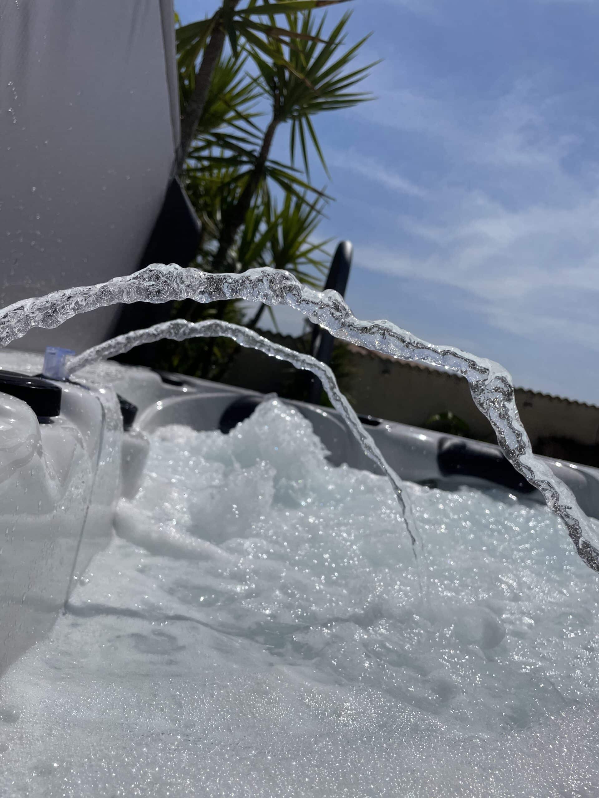 Jets d'eau épais et scintillants en arche au-dessus de la mousse bouillonnante d'un jacuzzi extérieur. Ciel bleu et palmiers.