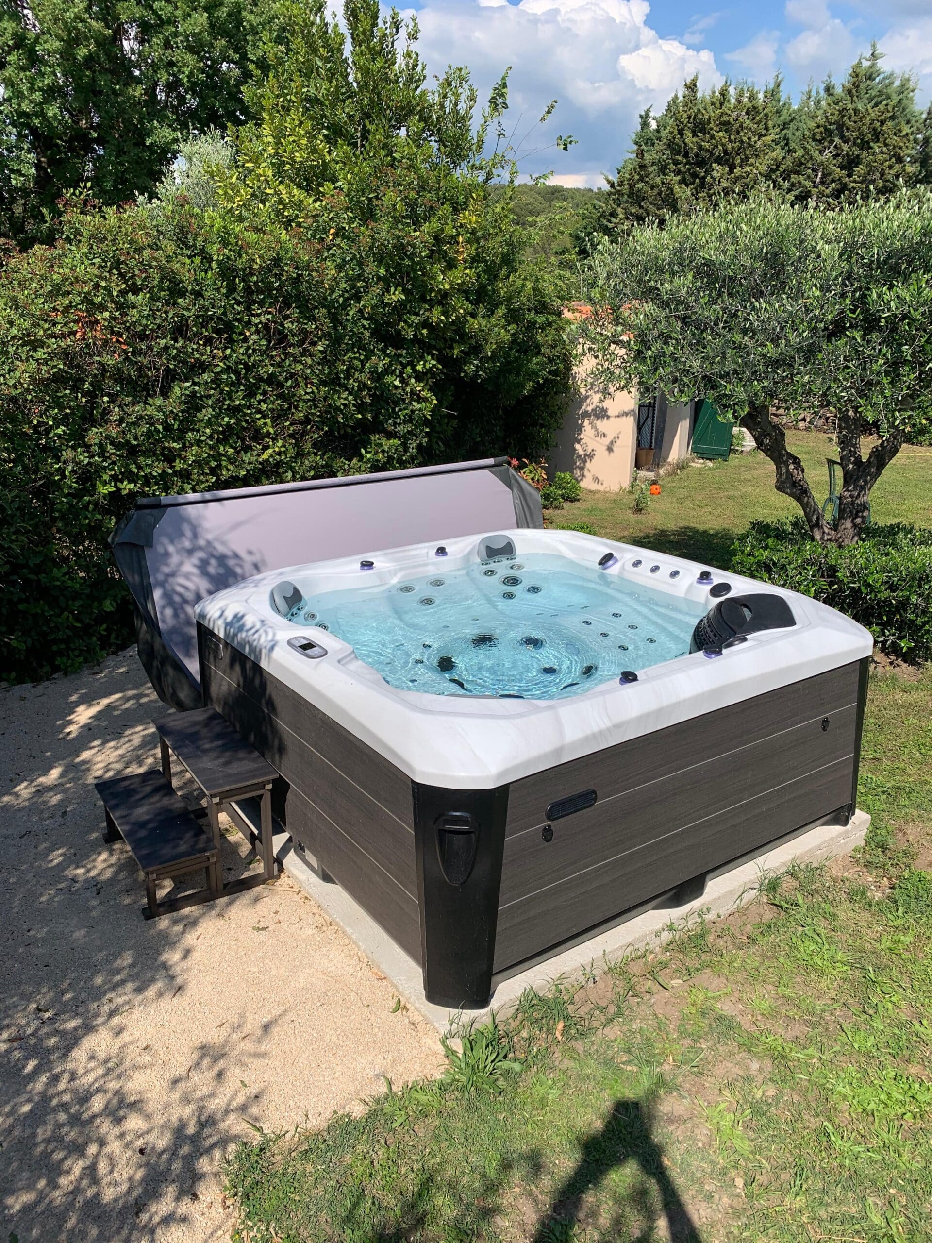 Spa jacuzzi extérieur moderne gris et blanc sur socle, prêt à la détente. Cadre luxuriant, soleil et olivier au bord du jardin.