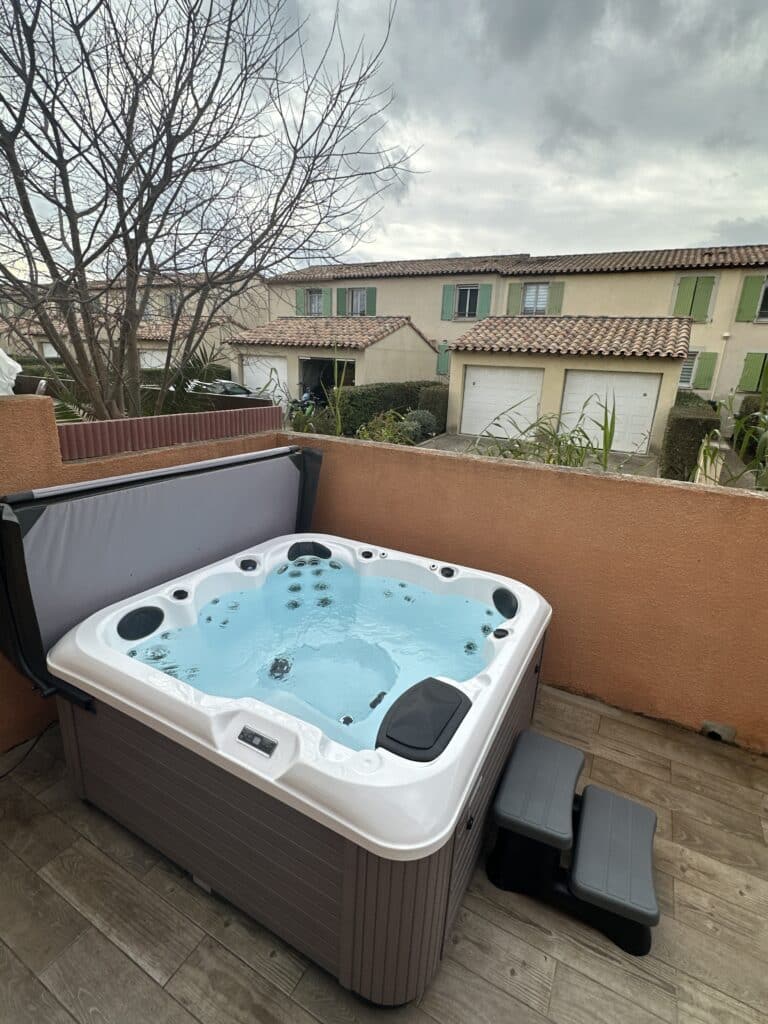 Grand jacuzzi extérieur moderne avec eau bleue bouillonnante sur une terrasse en bois, sous un ciel couvert, face à des maisons.
