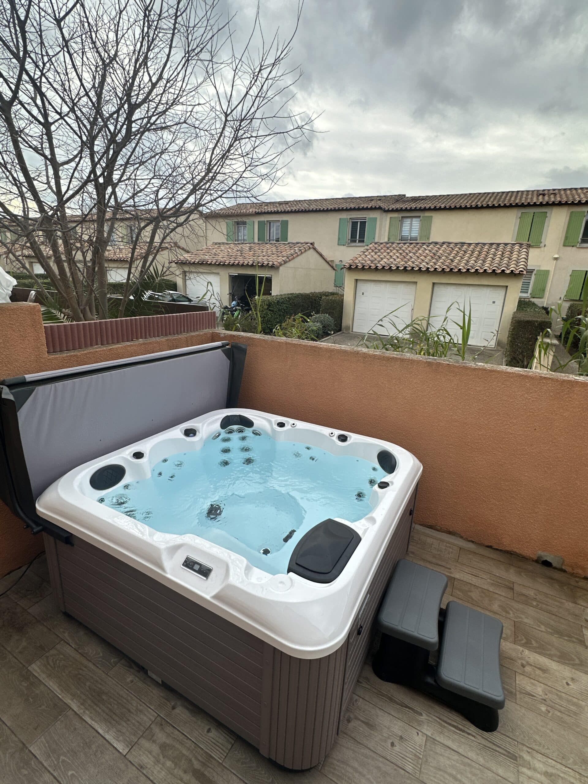 Grand jacuzzi extérieur moderne avec eau bleue bouillonnante sur une terrasse en bois, sous un ciel couvert, face à des maisons.