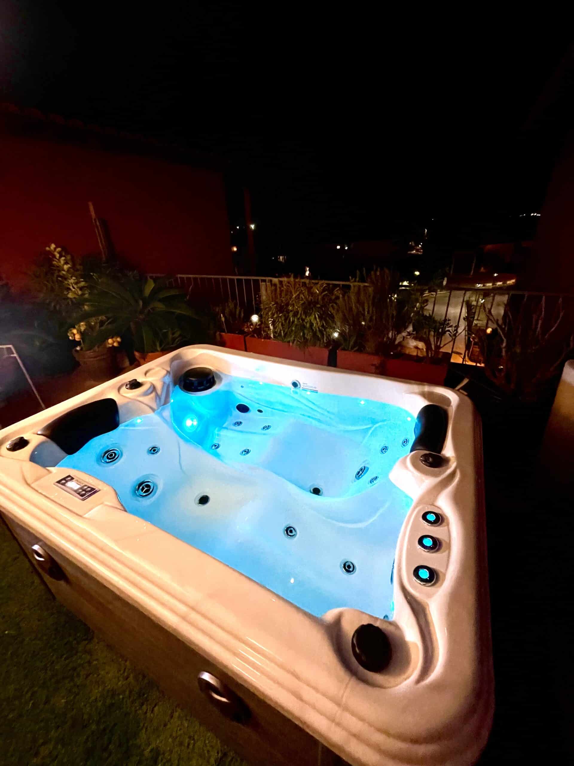 Jacuzzi extérieur sur terrasse la nuit. Éclairage LED bleu intense de l'eau, entouré de plantes et d'un mur rouge sombre, idéal pour la détente.