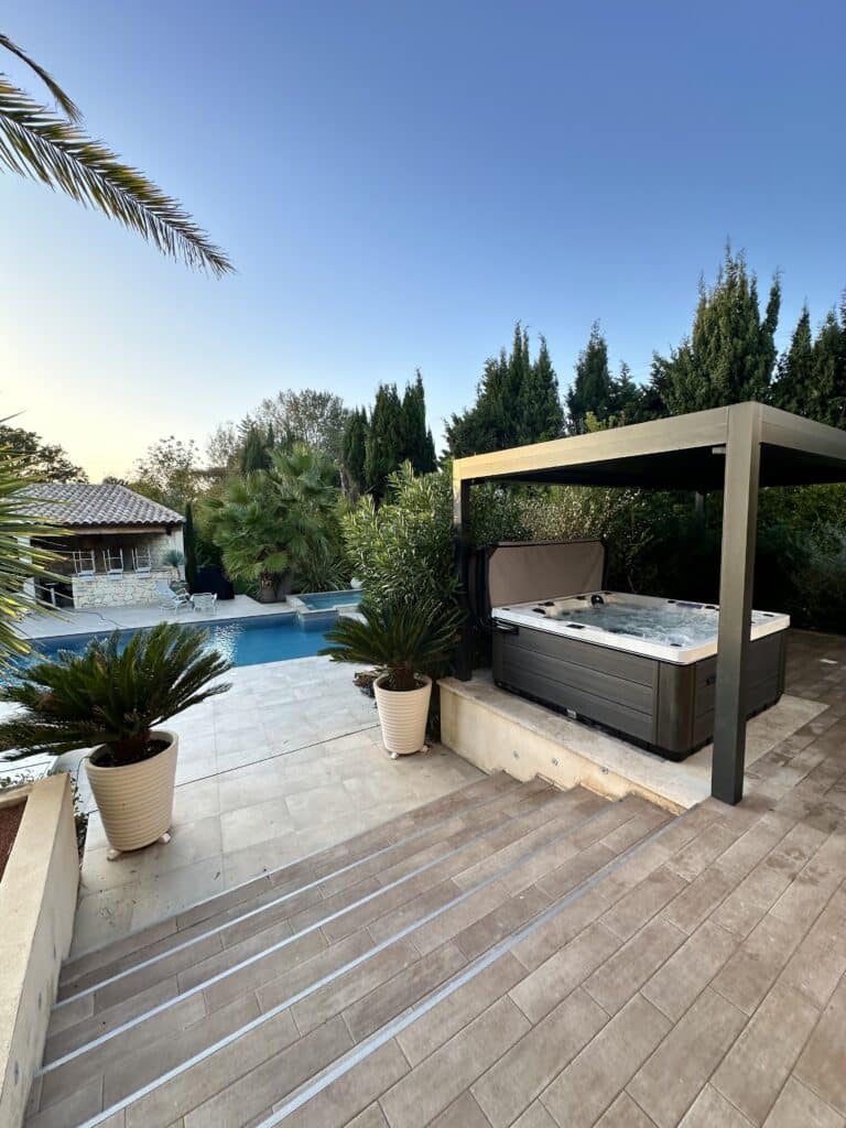 Jacuzzi sous pergola : design de terrasse et jardin. Jacuzzi gris sous pergola sur une terrasse en carrelage beige avec des marches. Piscine et cyprès luxuriants en arrière-plan.
