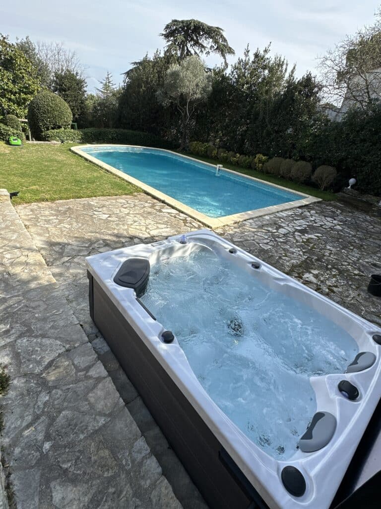 Jacuzzi et Piscine : Détente et Luxe au Cœur du Jardin Jacuzzi moderne bouillonnant sur une terrasse en pierre naturelle, à côté d'une longue piscine bleue dans un jardin privé.