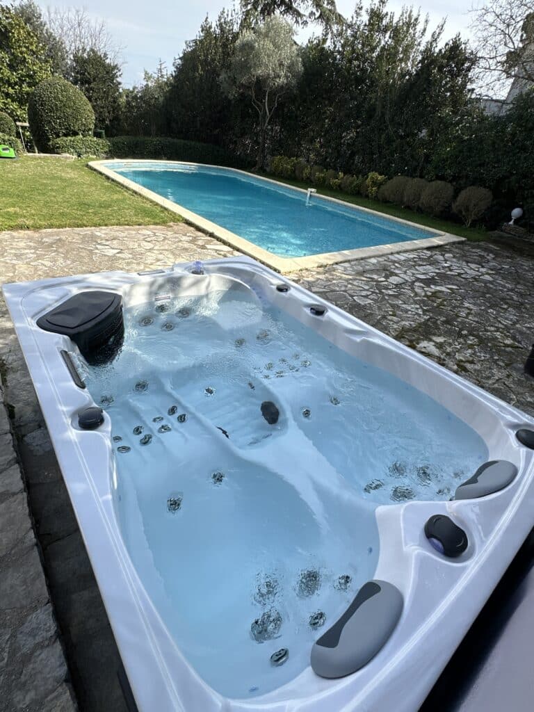 Jacuzzi extérieur blanc bouillonnant sur dalle de pierre rustique, devant une piscine rectangulaire bleue et un mur végétal dense.