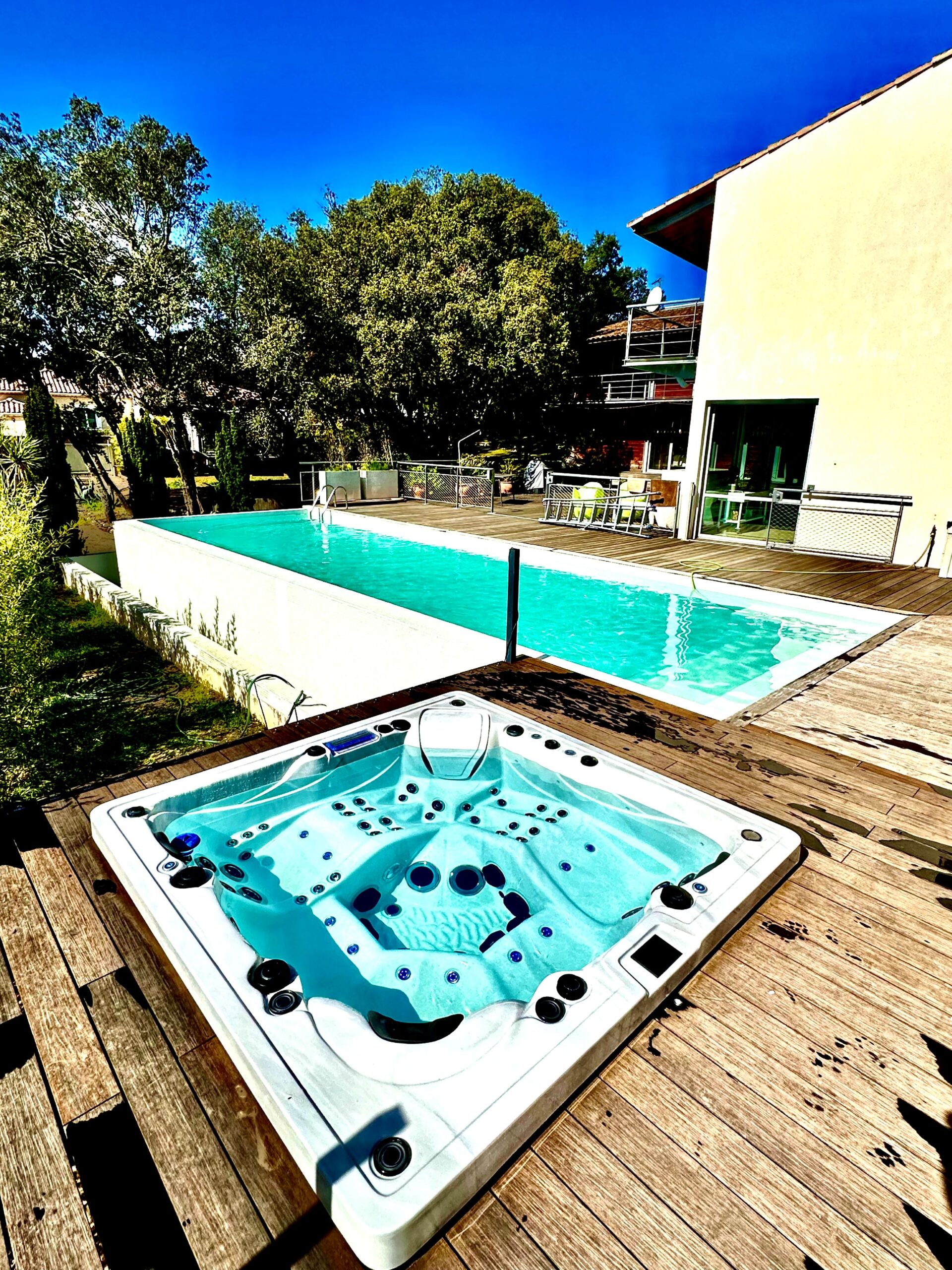 Luxueux jacuzzi extérieur et longue piscine turquoise sur une terrasse en bois. Villa moderne sous un ciel bleu éclatant et des arbres.