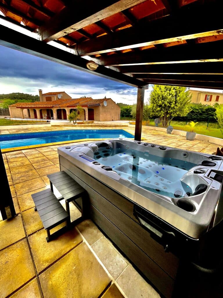 Jacuzzi, piscine, et villa provençale : luxe et détente. Luxueux jacuzzi sur une terrasse en dalles ocres, face à une grande piscine bleue et une villa provençale sous un ciel d'été orageux.