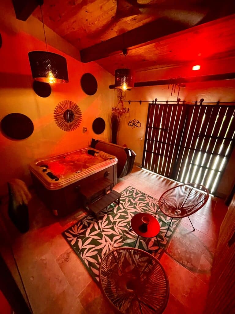 Jacuzzi privatif : Ambiance spa rouge pour une nuit romantique. Jacuzzi privé éclairé d'une ambiance rouge intense sous des poutres en bois. Fauteuils Acapulco et tapis végétal dans ce spa intérieur.