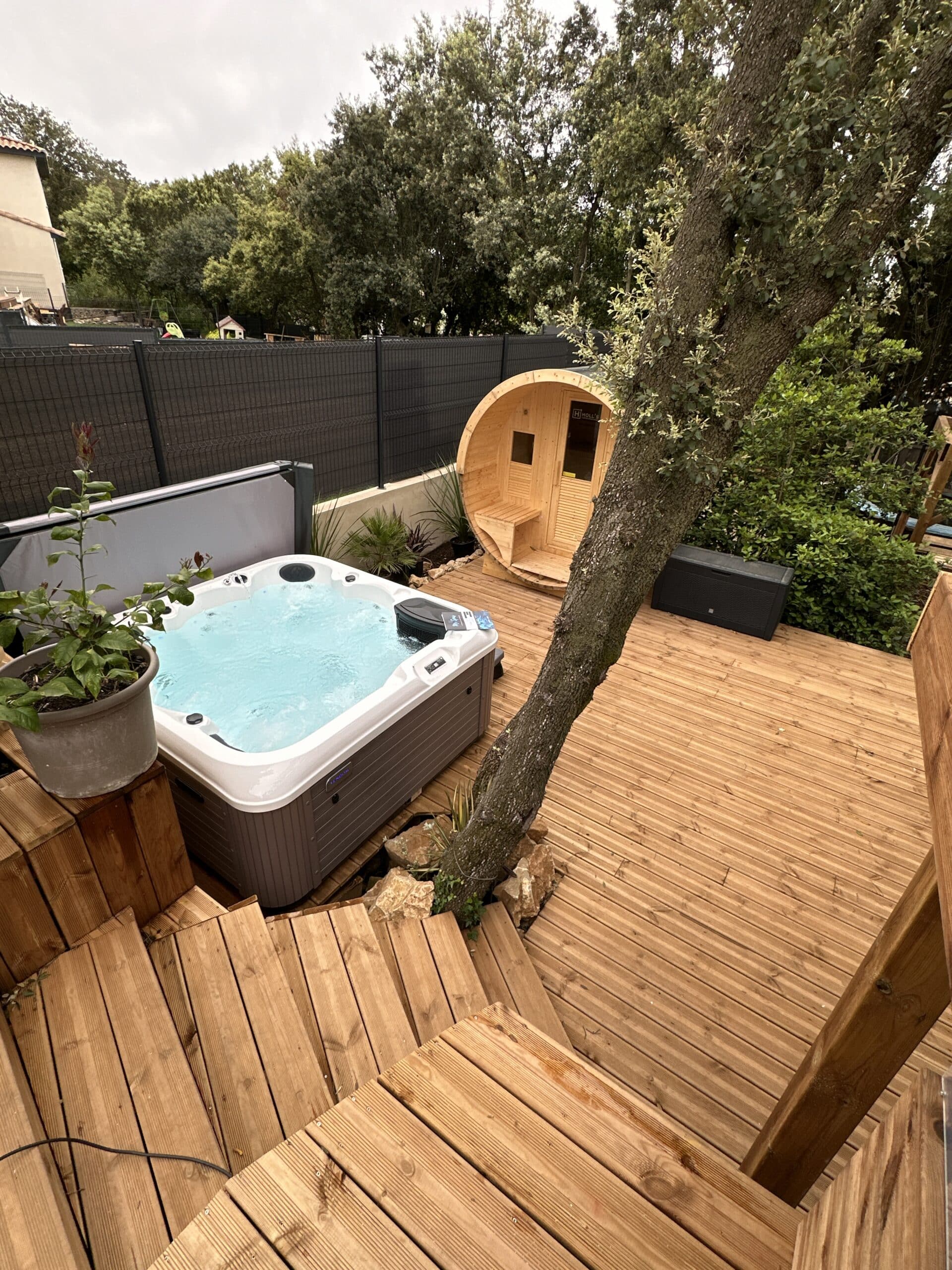 Aménagement extérieur luxueux : jacuzzi, sauna tonneau Holl's, et grande terrasse en bois, entourés d'un jardin luxuriant.
