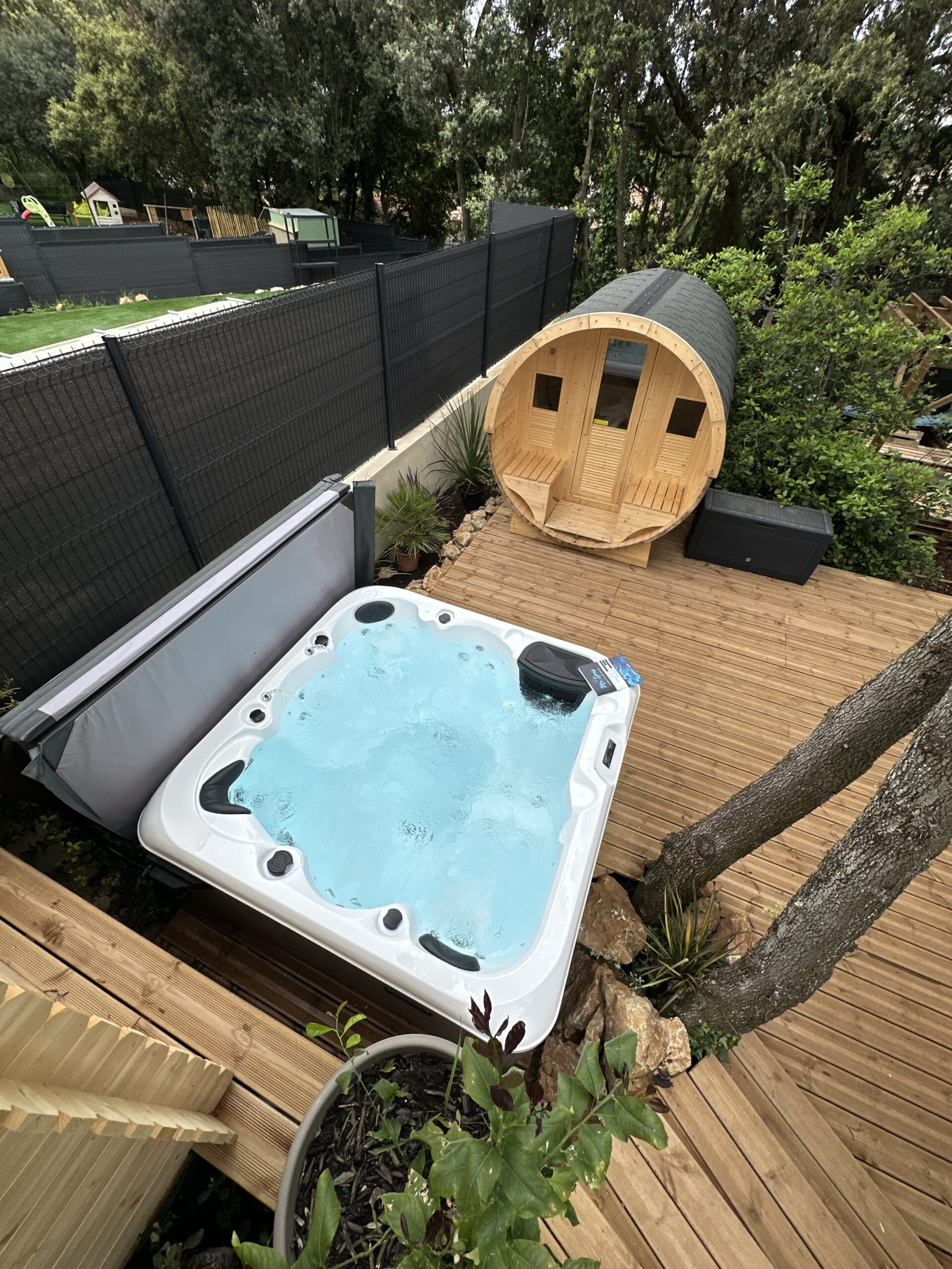 Terrasse en bois moderne avec jacuzzi extérieur bouillonnant et sauna tonneau en bois clair. Clôture noire et arbres luxuriants.