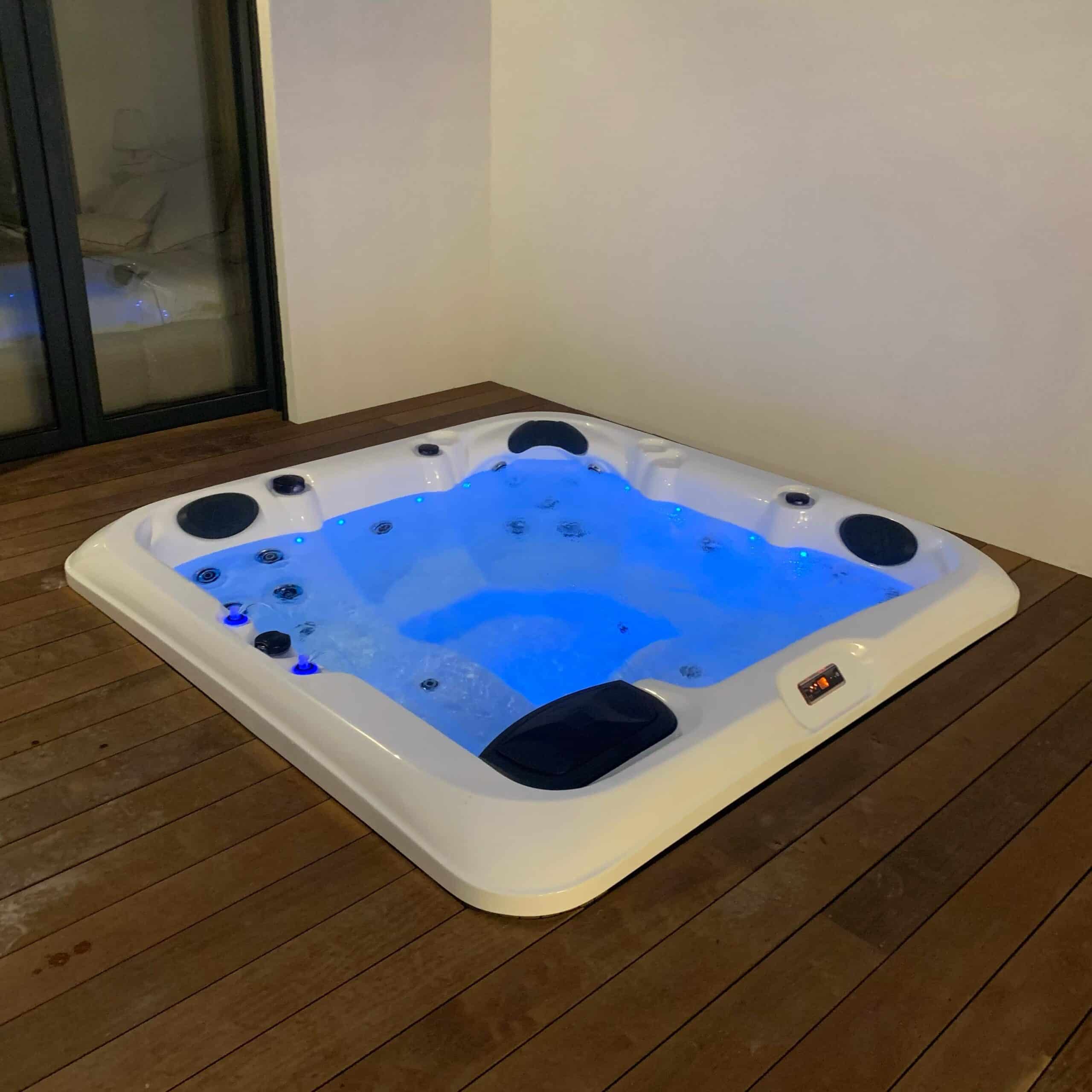 Jacuzzi d'extérieur blanc et moderne, encastré dans une terrasse en bois sombre. Le bain à remous brille d'une intense lumière LED bleue, invitant à la détente.