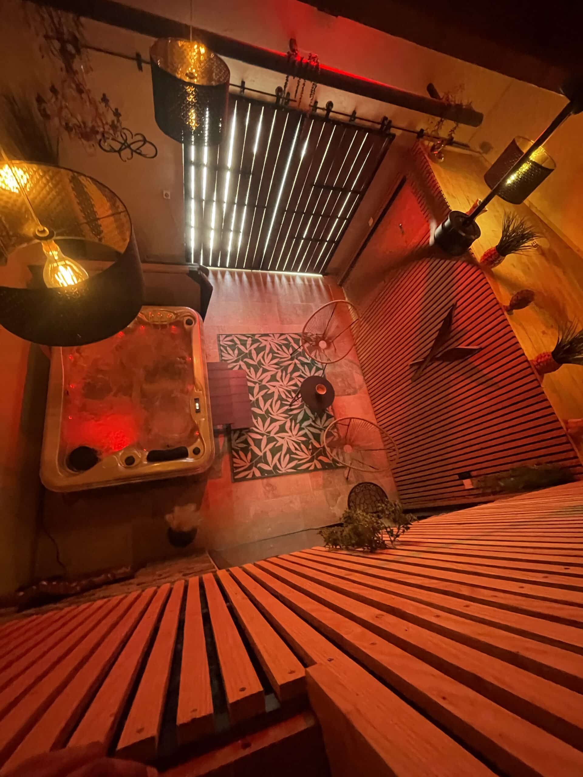Espace spa intimiste avec jacuzzi, éclairage ambre et rouge, sol en bois latté et tapis tropical. Ambiance chaleureuse.