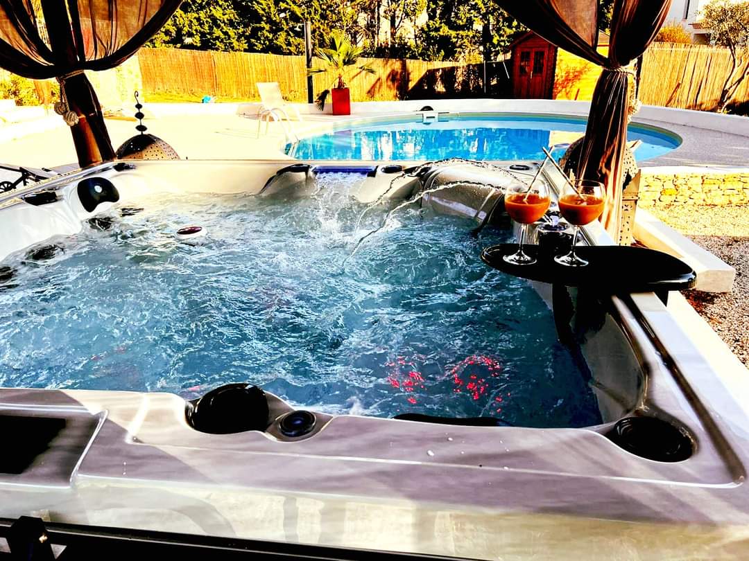 Spa extérieur luxueux avec jets d'eau, éclairage rouge, et deux cocktails. Piscine et palmiers en arrière-plan.