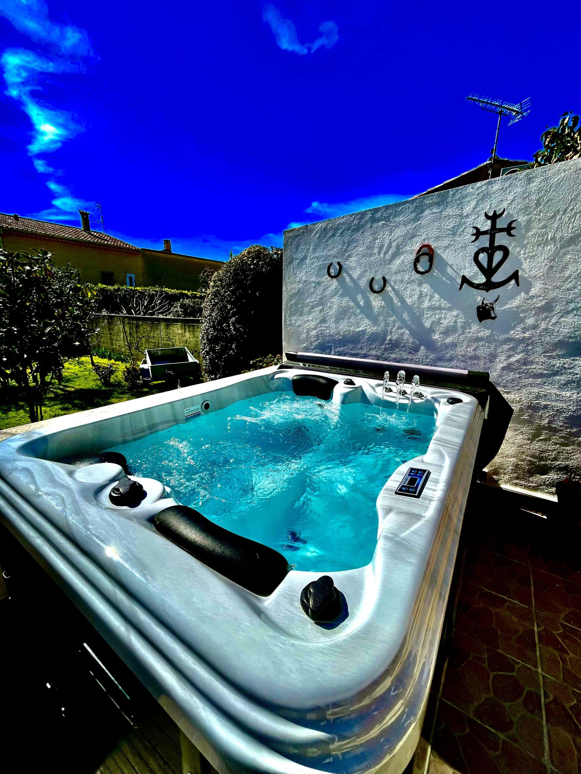 Jacuzzi extérieur bouillonnant, eau turquoise sous un ciel bleu profond. Mur blanc orné de l'ancre de Camargue, de fers à cheval et d'une tête de taureau.