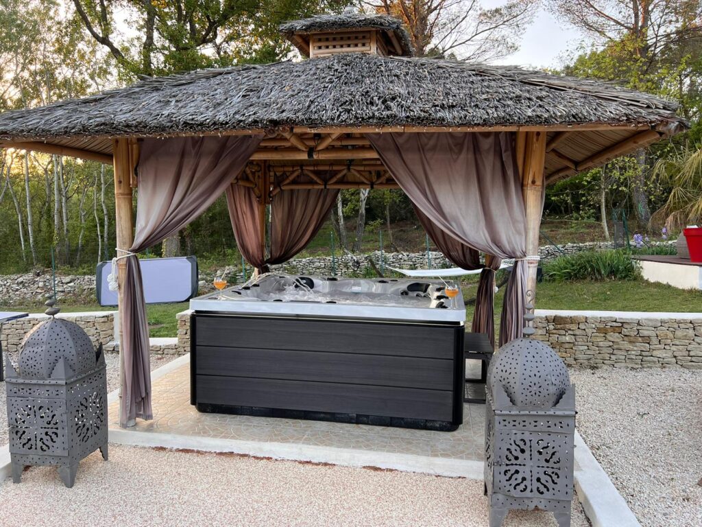 Spa extérieur de luxe : profitez du jacuzzi sous un gazebo exotique. Bain à remous gris sous gazebo en bambou au toit de chaume. Des rideaux et deux lanternes marocaines encadrent le spa extérieur luxueux.