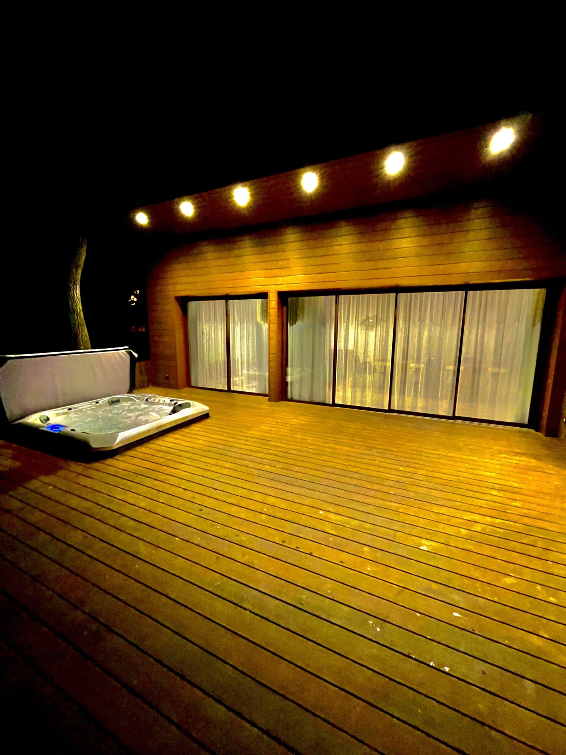 Jacuzzi moderne en fonction sur une grande terrasse en bois. Éclairage chaud de la façade en lattes et des baies vitrées voilées, nuit.