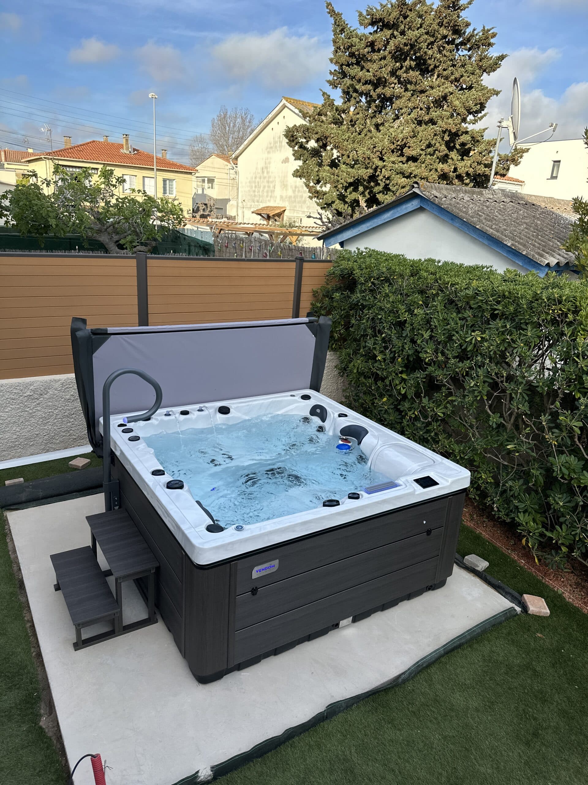 Jacuzzi spa Vendôm extérieur en fonctionnement, avec sa couverture grise ouverte et ses marches. Installation sur dalle, entouré de gazon et haie.