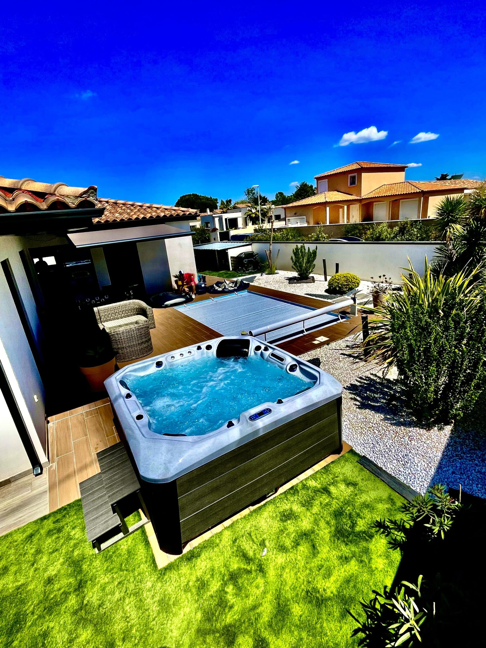 Grand jacuzzi extérieur bouillonnant sur une terrasse en bois luxueuse avec jardin et pelouse verte, sous un ciel bleu d'été.