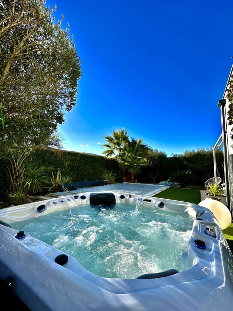 Jacuzzi spa en plein air : détente sous le ciel bleu éclatant. Jacuzzi spa bouillonnant dans un jardin luxuriant. Ciel bleu éclatant au-dessus d'oliviers et de palmiers. Ambiance relaxation.