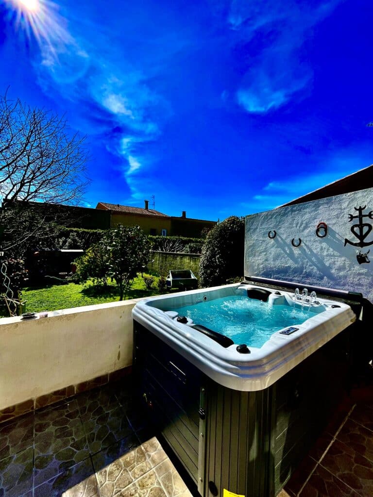 Spa Jacuzzi extérieur sous ciel bleu éclatant dans un jardin Jacuzzi bouillonnant sur terrasse ensoleillée, entouré d'un jardin. Ciel bleu profond, décor mural ancre et croix régionale.
