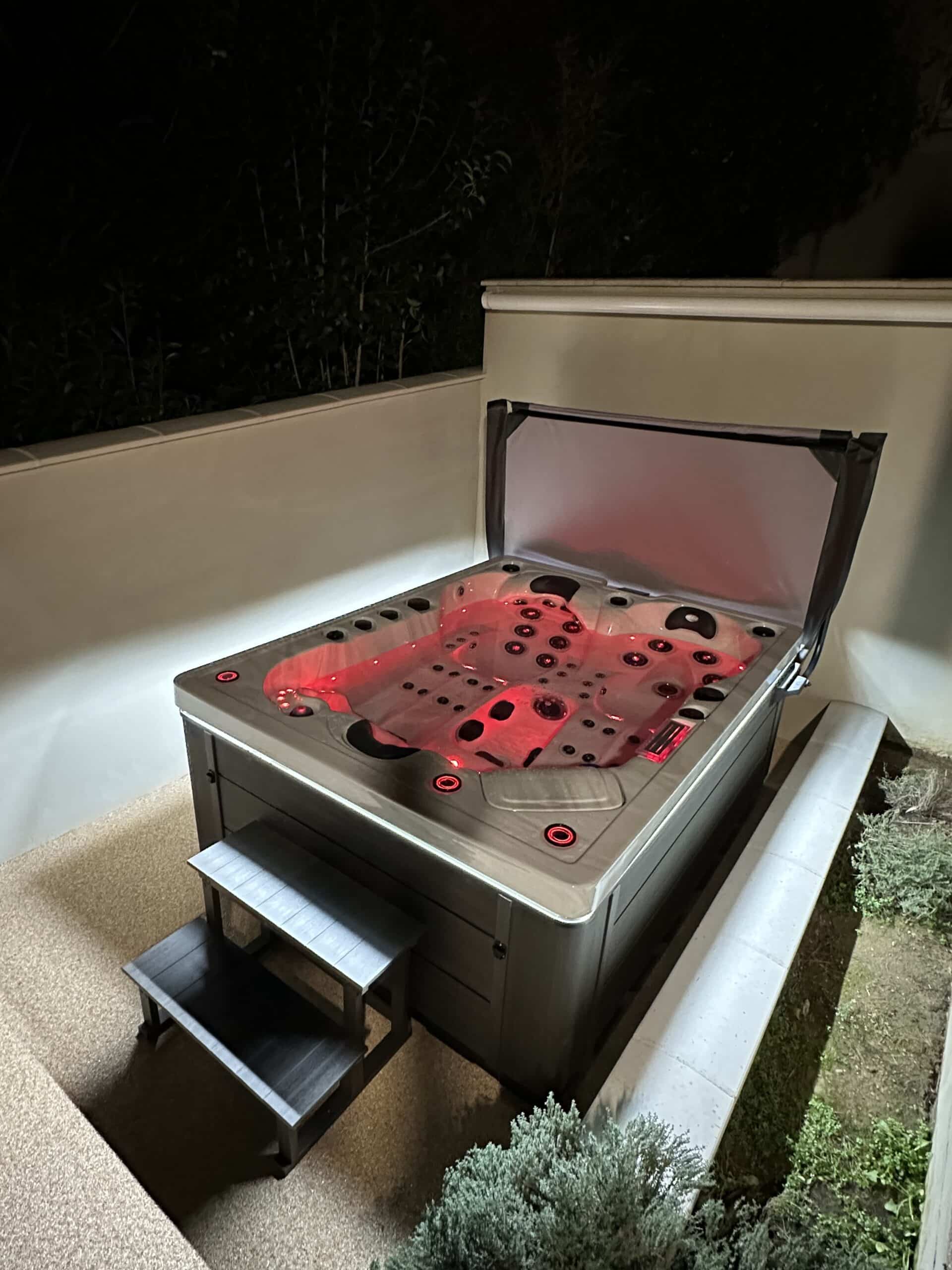 Spa extérieur moderne la nuit, cuve éclairée par des LED rouges vives. La couverture est levée et des marches grises sont installées.