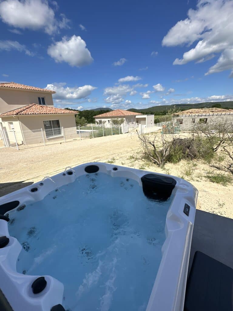 Jacuzzi extérieur moderne face aux maisons et montagnes. Jacuzzi extérieur blanc bouillonnant, avec jets d'eau, installé près de maisons neuves en construction. Beau ciel bleu et montagnes boisées.